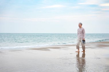Gülümseyen Asyalı son sınıf öğrencisi yalınayak ve elinde ayakkabılar, kumsalda yürümek, yaşlı yaşam tarzı, seyahat, doğada rahatlama.                             