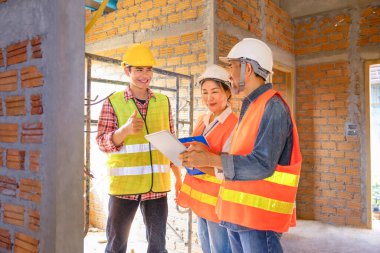 Genç Asyalı elektrikçi elektrik tesisatı hattını açıklıyor ve inşaat halindeki inşaat işçileri inşaat alanında müteahhit ekibine başparmak yapıyor. 