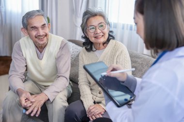 Asyalı yaşlı çift, tablet bilgisayar kullanan genç bir kadın sağlık personelini dinlemeye konsantre olup evdeki odada sağlık bilgileri veriyor.  