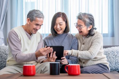 Mutlu Asyalı aile, yaşlı yetişkin baba ve anne ve genç yetişkin kızı koltukta oturuyor, gülümsüyor ve birlikte tablet bilgisayarına bakıyorlar, aile yaşam tarzı oturma odasında.  