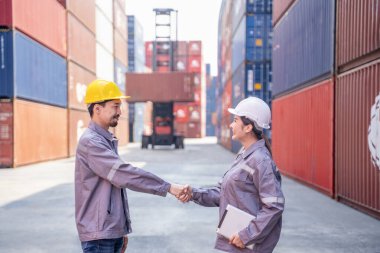 İş kıyafetleri ve güvenlik kaskı takan bir kadın ve erkek konteynır işçisi ticari limandaki konteynır kargosunu kontrol ettikten sonra el sıkışıyorlar.