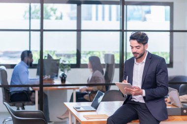 Tatilde ofis masasında oturan genç işadamının portresi dijital tablet, çok ırklı iş adamları, arka planda çalışan ofis çalışanları üzerinde çalışıyor.