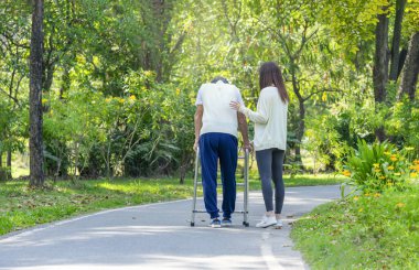 Kızının arka görünüşü engelli babaya parkta yürüme engelli yaşlı bir adamı destekleyen genç bir kadın parkta yürümek için yürüyen bir adamı destekliyor.