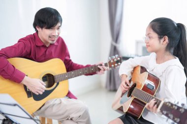 Odada öğretmenle gitar çalan Asyalı kız çocuğu, müzik öğrenmesine yardım eden yetişkin çocuk, müzik eğitimi kavramı, müzik eğitimi, klasik orkestra