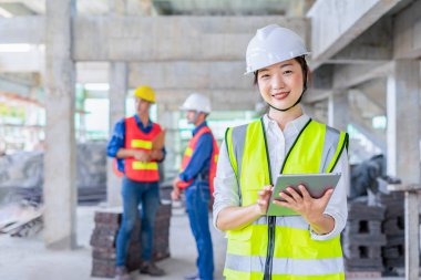 Asyalı kadın mühendis dijital tablet tutuyor, kameraya gülümsüyor ve poz veriyor, iş arkadaşları inşaat altında inceleme yapıyor.