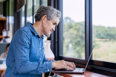 modern kıdemli profesyonel adam cam pencere kenarında oturuyor, işyeri sahibi dizüstü bilgisayara odaklanıyor