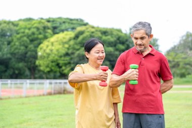 Asyalı son sınıf evli çift birlikte doğa parkında egzersiz yapıyor. Yaşlı adam, dambılla egzersiz yapmak için karısına destek oluyor.