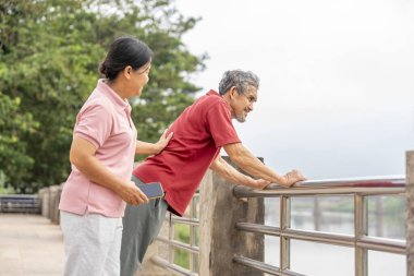 Sağlıklı yaşlı bir çift şehrin nehir kıyısında yürüyüş yolunda egzersiz yapıyor ve dinleniyor, yaşlı bir adam vücudunu esnetiyor.
