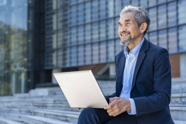 Yaşlı deneyimli iş adamı takım elbise giyiyor, dışarıda dizüstü bilgisayarla oturuyor, gülümsüyor ve ileriye bakıyor, başarıyı bekliyor.