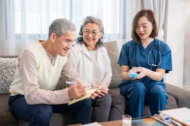 Asyalı kıdemli bir adam eşi için not alırken, konuk bir hemşire de ilaç alma, yaşlı sağlık hizmetleri, sağlık sigortası, evde sağlık hizmetleri gibi konularda tavsiye veriyor.