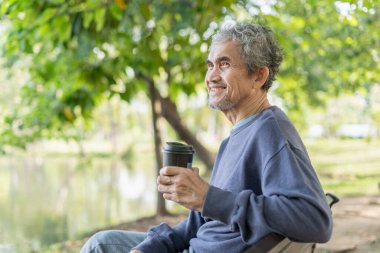 Portre gülümseyen Asyalı son sınıf öğrencisi. Doğa parkındaki bankta oturuyor. Elinde kahve fincanı, yaşlı yaşam tarzı, rahatlama.