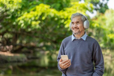 Gri saçlı ve sakallı mutlu bir adam, kulaklık takıyor, doğa parkında müzik dinlemek için akıllı telefon tutuyor, yaşlı yaşam tarzı, rahatlama.