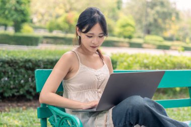 Genç ve güzel bir kadın dizüstü bilgisayar kullanıyor, parkta bankta oturuyor.