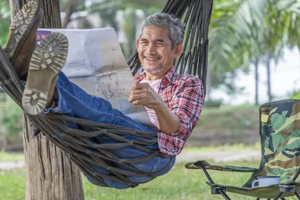 Yaşlı yaşam tarzı, seyahat ve kamp, mutlu kıdemli adam hamakta uzanıyor ve yazın bir kağıt haritaya bakıyor.