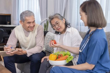 Üniformalı ev sağlık hemşiresi yaşlı çifte meyve, vitamin ve diyet liflerinin besin değerini açıklıyor.
