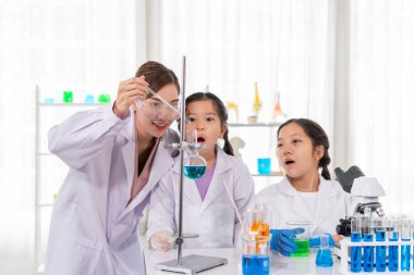 Genç bayan öğretmen, çocuğun mikroskopla, ilkokul sınıfıyla ve laboratuvar önlükleriyle fen bilgisi okuyan çocuklarla kimyasal deney yapmasına yardım ediyor.