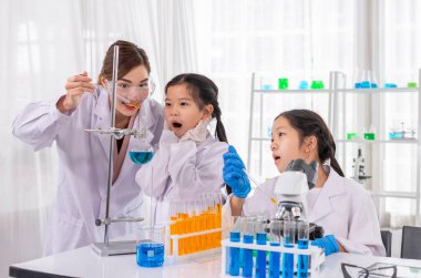 Genç bayan öğretmen, çocuğun mikroskopla, ilkokul sınıfıyla ve laboratuvar önlükleriyle fen bilgisi okuyan çocuklarla kimyasal deney yapmasına yardım ediyor.