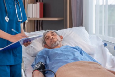 Hasta semptomlarını gözlemlemek için bir panoda tıbbi rapor tutan bir hemşireyle yatakta yatan kıdemli bir hastanın görüntüsü.