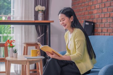 Genç ve çekici Asyalı kadın evdeki rahat bir odada kitap okumaya odaklan.