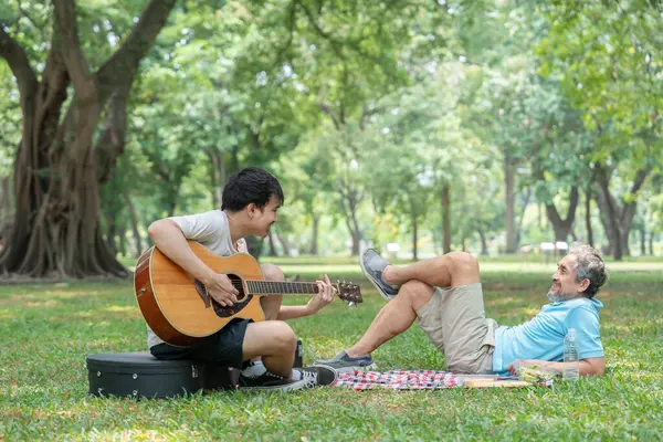 Büyük baba çimenlerin üzerinde yatıyor ve genç yetişkin oğul parkta gitar çalıyor, mutlu Asyalı aileyi birbirine bağlıyor tatilin tadını çıkarıyor, aile hayatı, ilişki.
