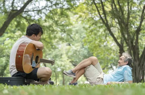 Büyük baba çimenlerin üzerinde yatıyor ve genç yetişkin oğul parkta gitar çalıyor, mutlu Asyalı aileyi birbirine bağlıyor tatilin tadını çıkarıyor, aile hayatı, ilişki.