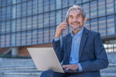 Üst düzey profesyonel iş adamı dizüstü bilgisayar yazmakla ve dışarıda otururken akıllı telefon kullanarak müşteriyle konuşmakla meşgul.