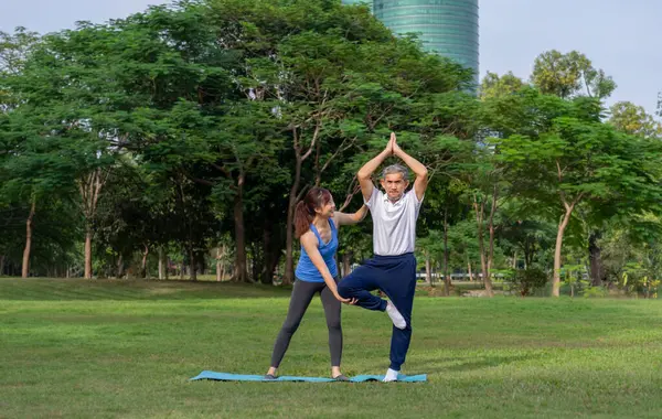 Genç yetişkin kız, yoga yapan yaşlı babaya yardım ve destek oluyor. Mutlu aile egzersizleri parkta, aile hayatı sporları konsepti. 