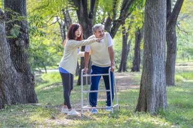Genç Asyalı kadın bakıcı yaşlı babaya yardım ediyor ve destek oluyor. Walker 'la parkta yürüyor, yaşlı rehabilitasyonu, sağlık hizmetleri...