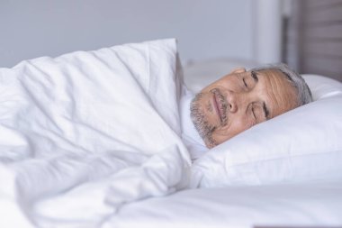 Üst düzey bir adam beyaz bir battaniyenin altında rahat bir şekilde yatıyor, dinleniyor, sıcak bir odada derin bir uyku çekiyor, yaşlılar yeterince uyuyor. 