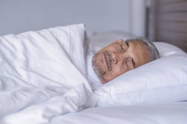 Üst düzey bir adam beyaz bir battaniyenin altında rahat bir şekilde yatıyor, dinleniyor, sıcak bir odada derin bir uyku çekiyor, yaşlılar yeterince uyuyor. 