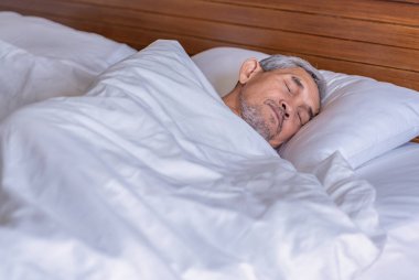 Üst düzey bir adam beyaz bir battaniyenin altında rahat bir şekilde yatıyor, dinleniyor, sıcak bir odada derin bir uyku çekiyor, yaşlılar yeterince uyuyor. 