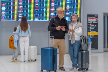 Asyalı yaşlı çift bavul ve sırt çantalı yolcular kalkış çizelgesinde bekliyor, uçağa binmek için akıllı telefondan çıkış ve yatış saatlerini kontrol edin, modern yaşam tarzına sahip yaşlılar, seyahat