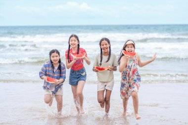 Karpuz tutan, sahilde eğlenen, deniz kıyısında birlikte koşan bir grup çok ırklı arkadaş.