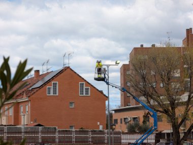 Bir işçi asansörde yükselir ve bir binaya ışıklandırma yerleştirilir. Etrafındaki yerleşim alanları ve ağaçlar bulutlu bir günde dağınık bulutlu bir zemin oluşturur..