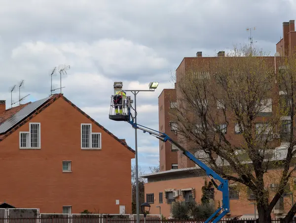 Bir işçi bir yerleşim yerindeki sokak lambasını tamir etmek için asansörle yükselir. Dekor tuğla binalar ve bulutlu bir gökyüzü içeriyor. Bu da öğleden sonra sahnesini gösteriyor..