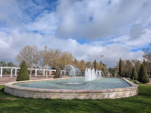 Dekoratif bir çeşme, öğleden sonra dinamik bulutlu gökyüzü altında düzgünce budanmış yeşillikler ve ağaçlarla çevriliyken dairesel bir gölette su sıçratır..