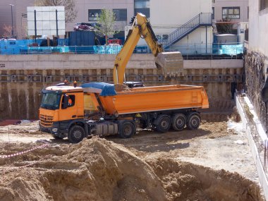 İşçiler, kentsel alandaki bir inşaat alanında toprak taşımak için turuncu bir çöp kamyonu ve kazıcı kullanıyorlar. Ağır makineler açık gökyüzü altında çalışırken kazı çalışmaları devam ediyor..