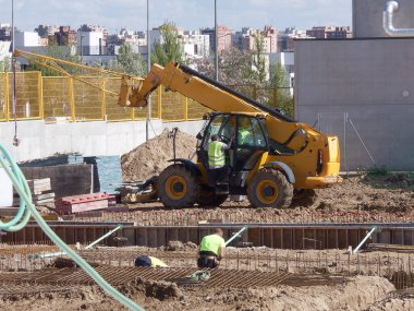 İki inşaat işçisi, açık gökyüzünün altındaki kentsel bir arazide ağır makineler ile bir inşaat sahasında görevlerini yerine getiriyorlar..
