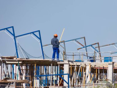 Bir inşaat işçisi, açık mavi gökyüzü altında yapısal elementlerin kurulumunu yöneten bir yapı iskeletinin üzerinde duruyor. Aletler ve iskeleler bölgeyi kuşatıyor..