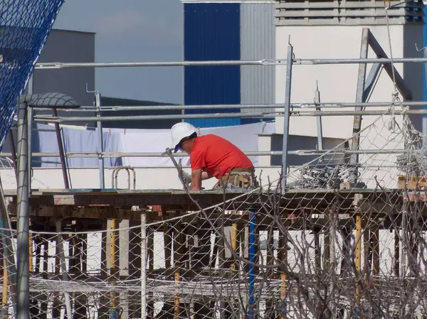 Kırmızı gömlekli ve beyaz kasklı bir işçi iskeleye eğiliyor ve inşaat alanındaki görevlere odaklanıyor. Ayar iyi aydınlatılmış ve düzenlenmiş..