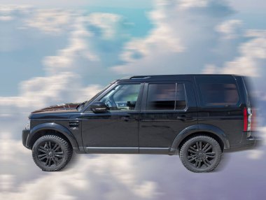 Siyah bir SUV, güneşli bir günde bulutlarla dolu bir gökyüzünde pürüzsüz bir şekilde dolaşarak mavi arkaplanda çarpıcı bir kontrast oluşturur. Sahne macera ve keşif duygusunu çağrıştırıyor..
