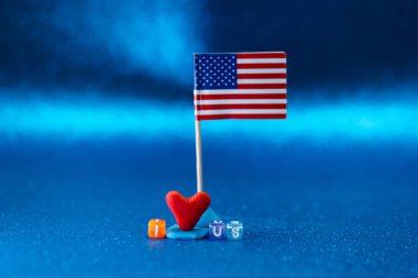 Plastikten yapılmış kırmızı bir kalbin yanında duran küçük bir Amerikan bayrağı. Renkli oyuncak harfleri parlak mavi bir yüzeyde 
