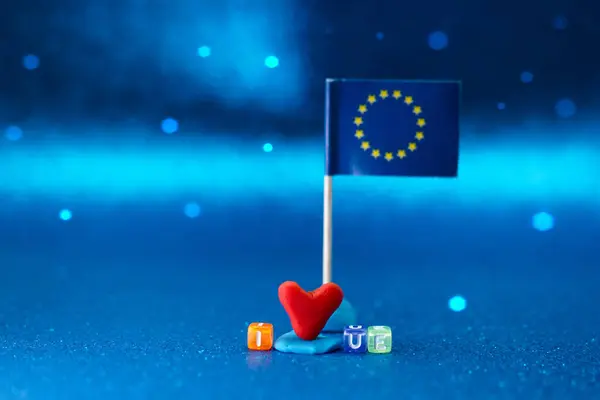 Çamurdan yapılmış küçük kırmızı bir kalp Avrupa Birliği 'ni temsil eden minyatür bir bayrağın yanında duruyor. Renkli harfler mavi tonlarla dolu bu neşeli ortamda destekleyici bir mesaj oluşturur.