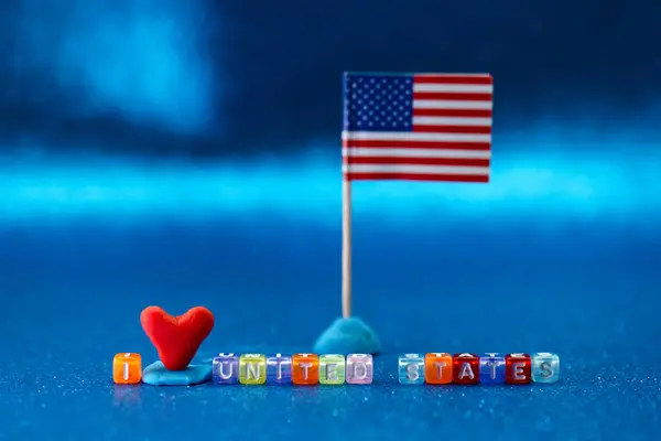 Küçük kırmızı bir kalp ve Amerika Birleşik Devletleri 'ni sevdiğimi söyleyen renkli boncuklar minyatür bir Amerikan bayrağının yanında oturuyor. Bu neşeli görüntü canlı mavi bir arkaplan üzerine kurulmuştur..