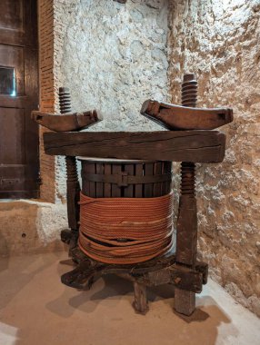 Kırsal bir taş mahzende, eski bir ahşap şarap presi belirgin bir şekilde duruyor ve geleneksel şarap yapımında kullanılan el yapımı araçları sergiliyor. Kalın ipler etrafını sarar ve gerçekliğini arttırır..