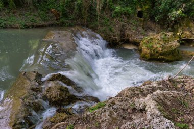 Su, canlı yeşil yapraklar ve davetkar doğal bir manzarayla çevrili kayalık bir çıkıntının üzerinden berrak bir havuza akar. Güneş ışığı sahneyi aydınlatır..