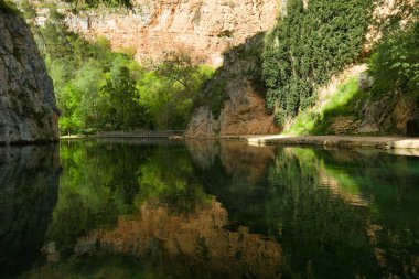 Kristal berrak su, canlı ağaçları ve sakin bir ortamda yükselen kayaları yansıtır. Tahta bir köprü çevrenin doğal güzelliğini arttırır..