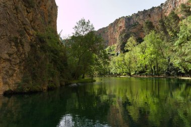 Sakin bir nehir, yüksek uçurumlar ve canlı yeşilliklerle çevrili yemyeşil bir vadiden yavaşça akar. Güneş ışığı ağaçların arasından süzülüp barışçıl bir atmosfer yaratır..