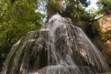 Kayalık bir uçurumdan dökülen su canlı yeşil ağaçlar ve parlak güneş ışığı altındaki bitki örtüsüyle çerçevelenmiş güzel bir şelale yaratarak barışçıl bir atmosferi çağrıştırıyor..