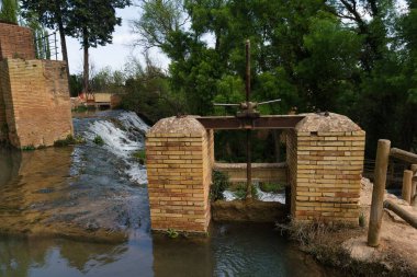 Etrafı yeşilliklerle çevrili eski bir baraj yapısının üzerinde çağlayan sular barışçıl bir atmosfer yaratıyor. Güneş akan suyun üzerinde parlıyor, sahnenin doğal güzelliğini arttırıyor..